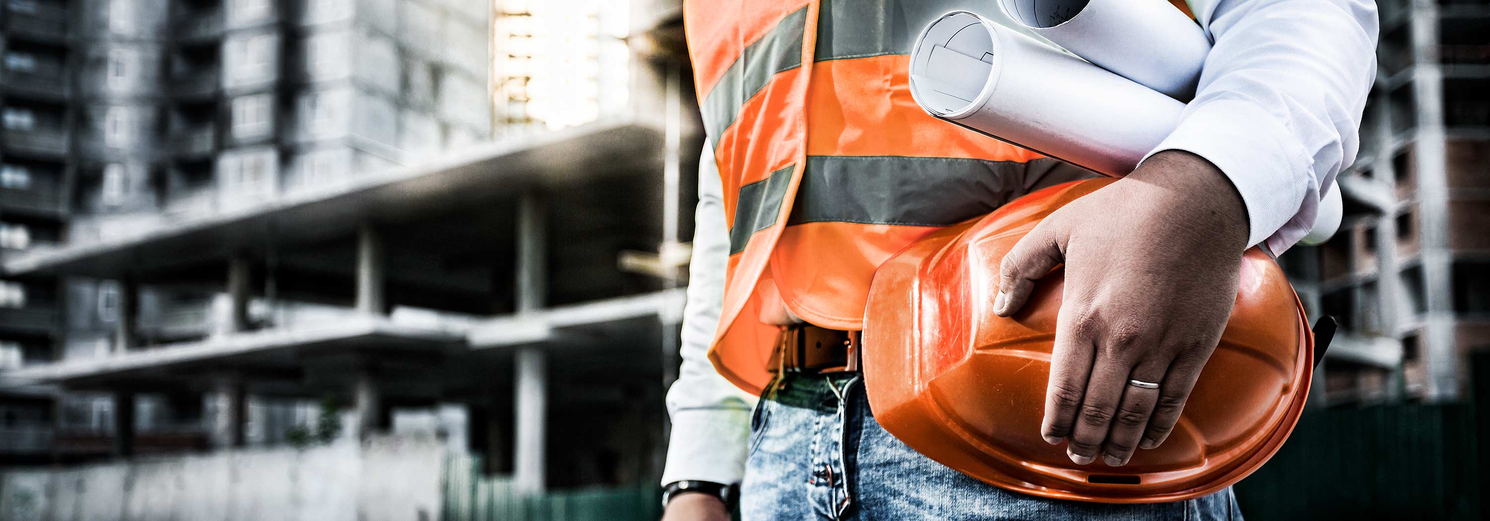 Titel Sliderbild von einem Bauarbeiter mit Bauhelm unter dem Arm vor dem Rohbau eines großen Gebäudes.